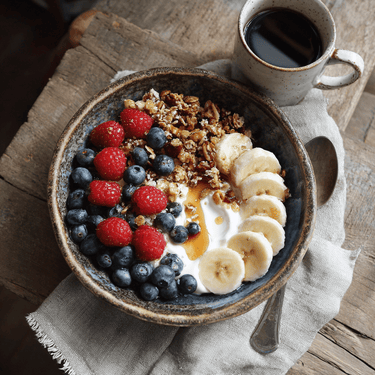 A rustic ceramic bowl with yogurt, blueberries, raspberries, banana slices, My Sister’s Granola – Nutty, Sweet, and Totally Addictive, toasted coconut, and honey sits beside a cup of black coffee on a wooden table.